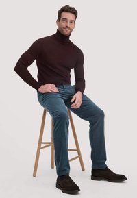 Burgunderfarbener Rollkragenpullover, blaue Cordhosen und dunkelbraune Wildlederschuhe. Modell sitzt auf einem Holzstuhl vor einem schlichten Hintergrund.