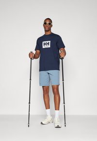 Tumesinine T-särk valge logoga, helesinised lühikesed püksid, valged sokid, beežid sneakersid ja mustad matkakepid. Mudel kannab prille.