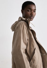 Beige, könnyű kapucnis dzseki cipzárral elöl, rugalmas húzózsinórral és oldalsó zsebekkel. Simább textúra, laza dizájn.