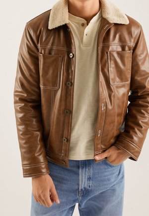 Man draagt een bruine leren jas met shearlingkraag, beige henley-shirt en blauwe spijkerbroek, met één hand in de zak.