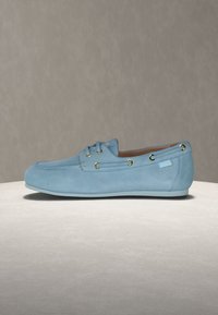 Γαλάζιο μοκασίνι από σουέντ με χρυσά μεταλλικά τρουκ, δέσιμο στο πλάι και επίπεδη σόλα, τοποθετημένο σε ανοιχτό γκρι επιφάνεια πάνω σε ουδέτερο φόντο.