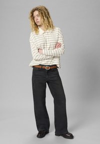 Mlada žena s dugom kovrčavom plavom kosom stoji s prekriženim rukama, obučena u kremasto prugastu košulju dugih rukava, crne traperice i smeđi remen.