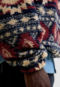 Sudadera de forro polar con una textura estampada en colores rojo, azul marino y crema. Presenta un corte holgado y puños de canalé, con una capa de denim por debajo.