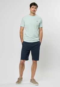 Jasnozielona koszulka z krótkim rękawem z małym logo, zestawiona z granatowymi szortami i beżowymi sneakersami, charakteryzująca się luźnym krojem i gładką fakturą.