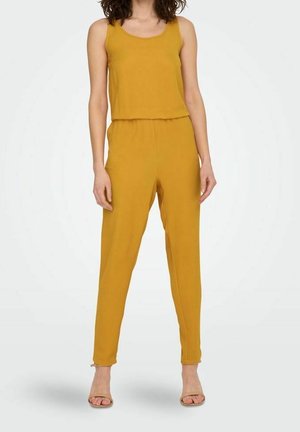 Musztardowy kombinezon z designem na ramiączkach i elastycznym paskiem w talii. Gładka tekstura, zwężane nogawki i długość do kostek. Klasyczny styl obuwia.