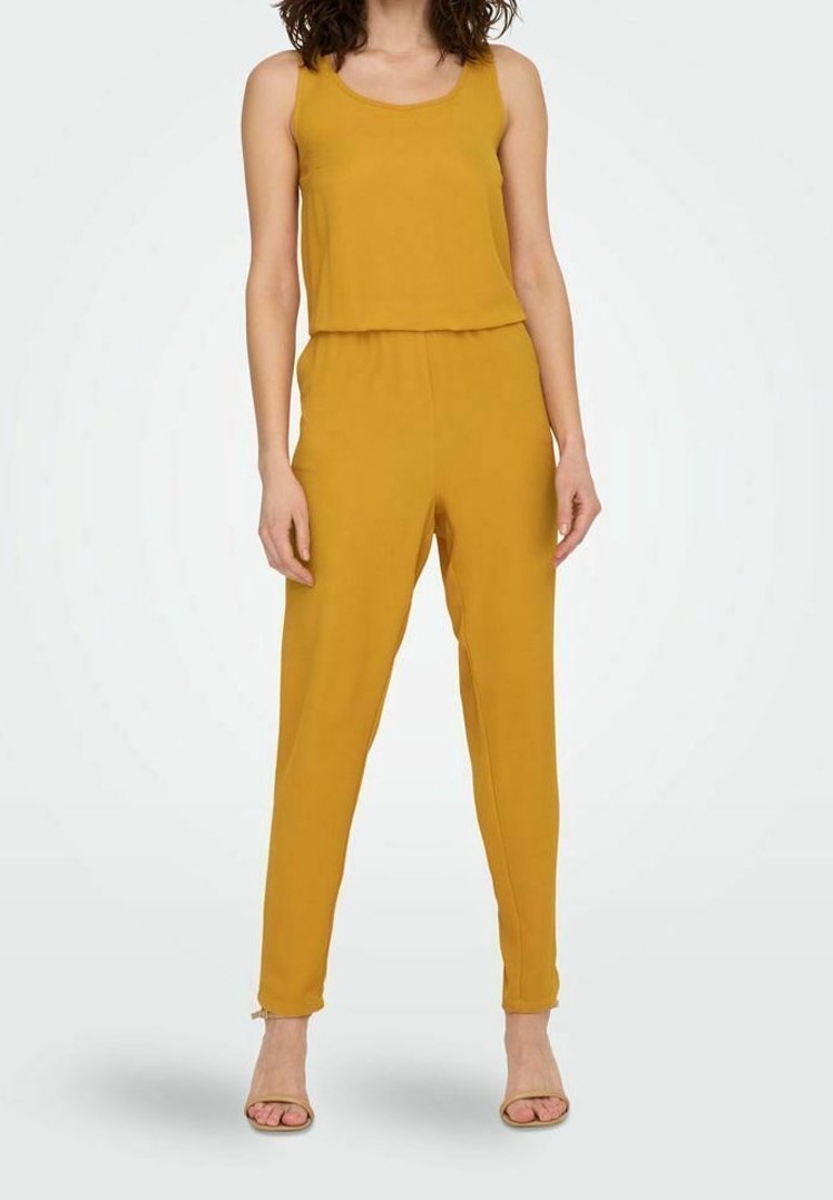 Combinaison jaune moutarde avec un design de débardeur et une ceinture élastique. Texture lisse, jambes fuselées et coupe à la cheville. Chaussures de style classique.