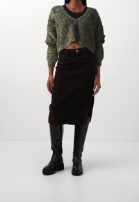 Pull court vert texturé avec col en V, assorti à une jupe midi en velours côtelé marron avec une fente latérale, et des bottes noires montant jusqu'aux genoux.