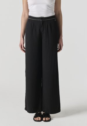 Mujer vistiendo pantalones negros de pierna ancha y cintura alta con pliegues, una camiseta blanca sin mangas metida por dentro y sandalias negras abiertas en la punta.