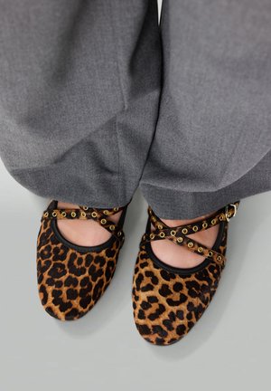 Leopardprint ballerina's van zacht materiaal met zwarte bies, met kruislings banden versierd met goudkleurige ringen. Grijze stoffen broek zichtbaar.