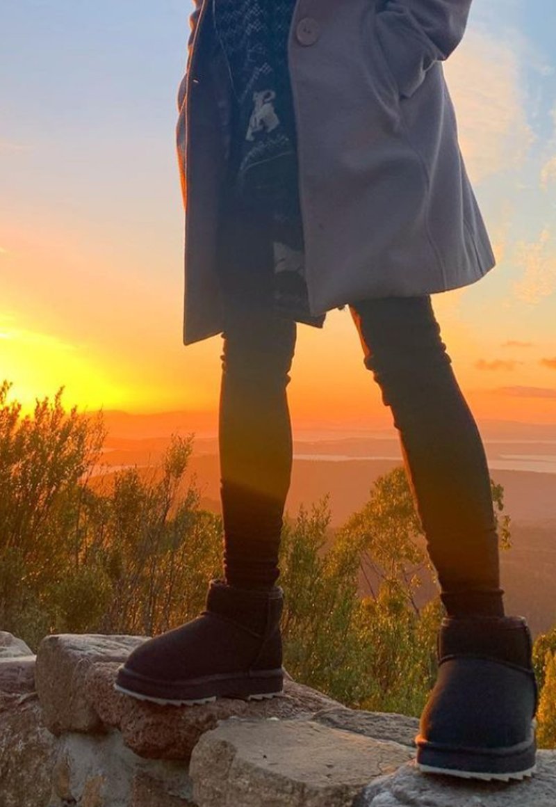 Czarne botki za kostkę o miękkiej powierzchni, z grubą podeszwą i zawiniętym mankietem, noszone z ciemnymi dopasowanymi spodniami na tle zachodu słońca.