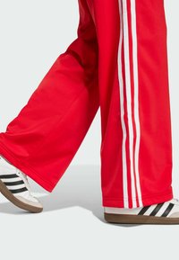 Red athletic pants with white side stripes, paired with white shoes featuring black stripes and brown rubber soles. Plain background.
