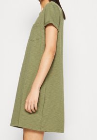 Robe olive verte à manches courtes avec une coupe décontractée, dotée d'une poche poitrine et d'un tissu doux et texturé. Design décontracté arrivant au genou.