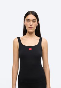 Μαύρο αμάνικο τοπ με ριμπ υφή, φαρδιές τιράντες και ορθογώνιο κόκκινο λογότυπο στο κέντρο. Απλός σχεδιασμός, εφαρμοστή γραμμή και λείο ύφασμα.