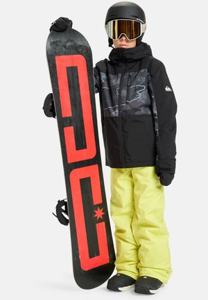 Veste de snowboard noire avec des accents gris, pantalon jaune fluo, et un snowboard noir portant un logo rouge. Des lunettes de neige complètent la tenue.