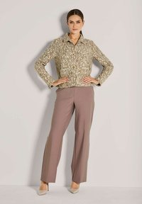 Frau steht mit den Händen in die Hüften gestützt, trägt eine gemusterte beige Jacke, mauve gerade geschnittene Hose und beige spitze High Heels.