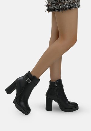 Gambe che indossano collant nude trasparenti e stivaletti in pelle nera con tacco largo, cerniere laterali e cinturini con fibbia.