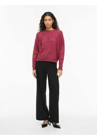 Pull en tricot magenta avec broderie florale sur la poitrine, assorti à un pantalon noir taille haute à jambes larges et des chaussures noires à talons.