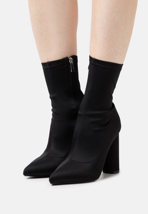 Botas de satén negro hasta el tobillo con una punta afilada, un ajuste ceñido y un cierre de cremallera en el lateral, que presentan un tacón alto y grueso.