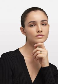 Gouden ringen met ingewikkelde designs gedragen aan vingers, model in een zwart geribbeld V-hals top, neutrale achtergrond benadrukt de gelaatskenmerken.