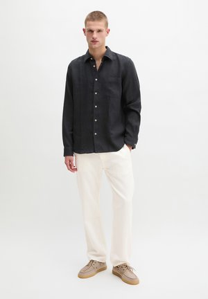 Nuori mies seisoo päällään musta napillinen paita, valkoiset housut ja beigea nauhalliset kengät yksinkertaisen valkoisen taustan edessä.