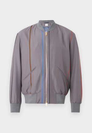 Göndör hajú férfi szürke bomber dzsekit, fekete pulóvert, világoskék bőszárú farmert, fekete cipőt és fekete övet visel, egyik kezével a zsebében áll.