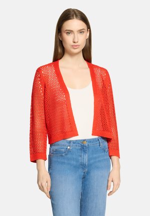 Femme portant un cardigan rouge vif à mailles ajourées sur un haut blanc et un jean taille haute bleu clair, debout devant un fond uni.