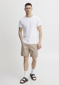 Weißes Baumwoll-T-Shirt, beiges hochgekrempeltes Shorts, weiße Socken, braune Sandalen; Model steht vor einem neutralen Hintergrund, die Hände in den Taschen.