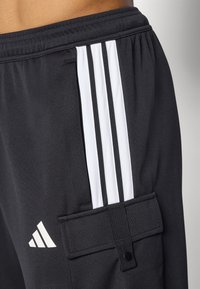 Black athletic pants with white three-stripe design and an adidas logo. Features a textured fabric and a side pocket with a snap closure.