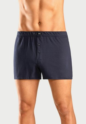 Männlicher Oberkörper und Oberschenkel, die dunkelblauen Baumwollboxershorts mit zwei vorderen Knöpfen und elastischem Bund tragen, vor einem schlichten Hintergrund.