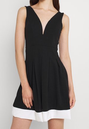 Sort ærmeløs kjole med dyb V-hals, plisseret design og en kontrasterende hvid kant. Fremstillet af glat stof, tætsiddende ved taljen.