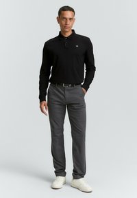 Camisa polo negra de manga larga con cuello y logo, combinada con pantalones grises y zapatillas blancas. La tela parece suave y ajustada.