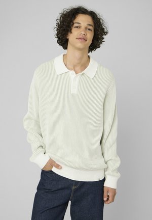 Jeune homme aux cheveux bouclés portant un pull en maille texturé clair avec un col et un jean bleu foncé, debout avec une main dans la poche.