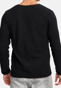 Chemise noire à texture, à manches longues, avec un col rond et des poignets ajustés, associée à un pantalon gris clair, vue de dos.