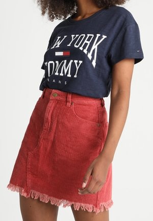 Vrouw draagt een marineblauw T-shirt met de tekst "New York Tommy Jeans" in de taille gestopt, gecombineerd met een hoog-laag rode corduroy rok met rafelige zoom.