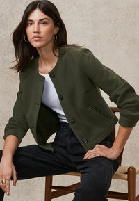 Olive green cropped jacket with a round neckline, black buttons, and two front pockets, paired with a white top and black pants.