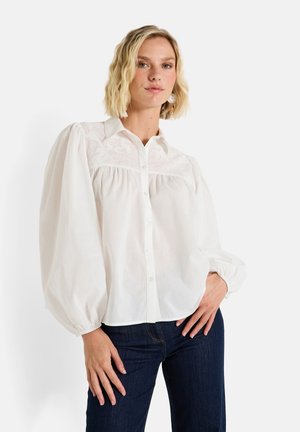 Chemise blanche à manches longues bouffantes, avec des broderies sur le plastron et en tissu doux. Portée avec un jean bleu foncé.