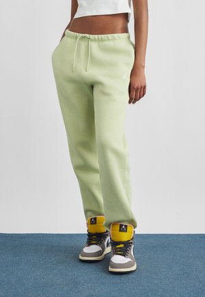 Osoba v svetlo zelených teplákoch s rukami v vreckách, bielom krátkom topom a sivobielozlatých vysokých teniskách na modrom koberci.