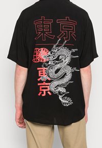 Maglietta nera a maniche corte con una grafica di drago bianco e testo rosso sul retro. Il tessuto appare morbido con una vestibilità comoda.