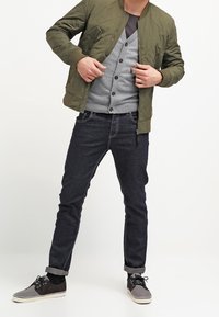 Olivově zelená prošívaná bomber bunda přes šedý cardigan s knoflíky, spárovaná s tmavými džíny a černými šněrovacími teniskami se šedými akcenty.