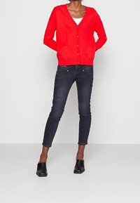 Czerwony dziergany kardigan z dekoltem w serek i zapięciem na guziki, zestawiony z ciemnymi obcisłymi dżinsami typu skinny i czarnymi klapkami na słupku.