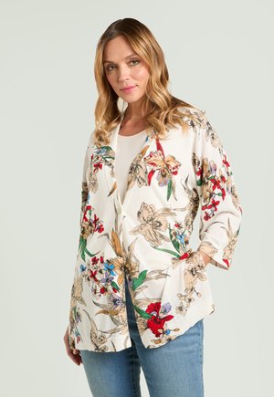 Kvinde med bølget blondt hår iført en hvid cardigan med blomstermønster og knapper over en hvid top og blå jeans, poserer foran en ensfarvet baggrund.