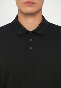 Juoda polo marškinėliai su tekstūruotu apykaklė, trijų mygtukų užsegimu ir siuvinėtu logotipu ant krūtinės. Trumpomis rankovėmis, klasikinio silueto.