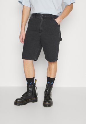 Persona que lleva pantalones cortos de denim negro, botas negras altas y calcetines negros con estampados de dinosaurios azules, de pie frente a un fondo liso.