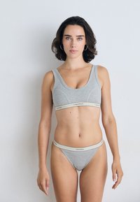 Vrouw die tegen een effen muur staat, gekleed in een grijze Calvin Klein sportbeha en bijpassende onderbroek, met een neutrale uitdrukking recht in de camera kijkend.