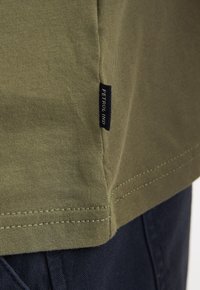 Camiseta de algodón color verde oliva con una etiqueta en la costura lateral, costuras en contraste y un ajuste relajado. El tejido tiene una textura suave y lisa.