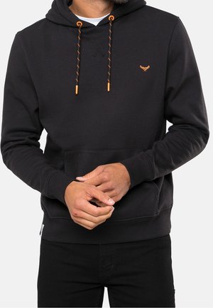 Homme portant un sweat à capuche noir avec des cordons de serrage orange et un petit emblème orange, les mains jointes devant, sur un fond clair.
