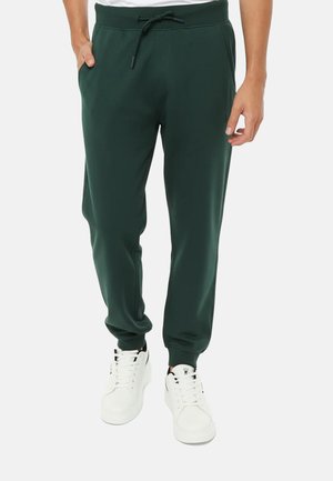 Uomo con pantaloni jogger verde scuro con coulisse in vita e sneakers bianche, in piedi con una mano in tasca.