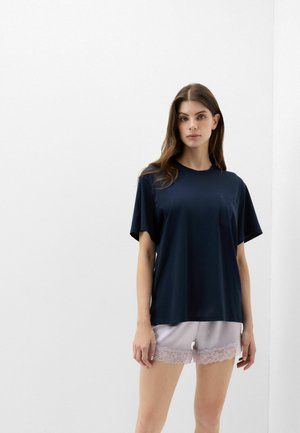 Vrouw draagt een loszittend marineblauw T-shirt en lichtroze korte broek met kant, staand tegen een witte muur.