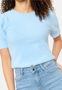 Světle modrý krátký svetr s krátkým rukávem vyrobený z měkkého a nadýchaného materiálu. Má kulatý výstřih a volný střih, kombinovaný s džíny s vysokým pasem.