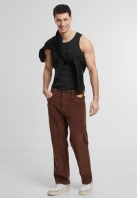 Brown corduroy pants with a loose fit, paired with a black tank top and a black sweater draped around the shoulders. White sneakers complete the look.
